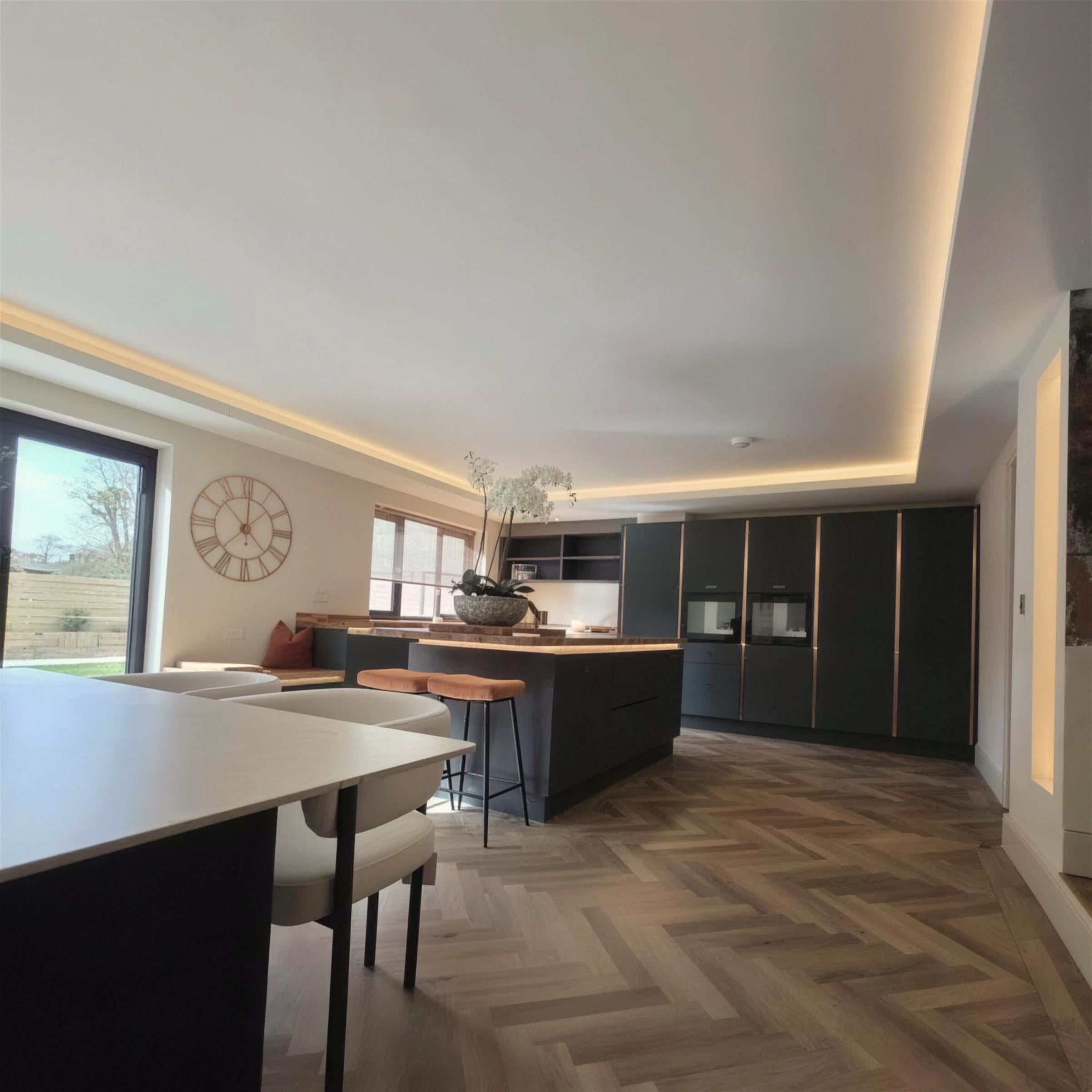 Open-plan kitchen with warm LED cove lighting running the full ceiling perimeter above dark cabinetry and herringbone oak flooring.