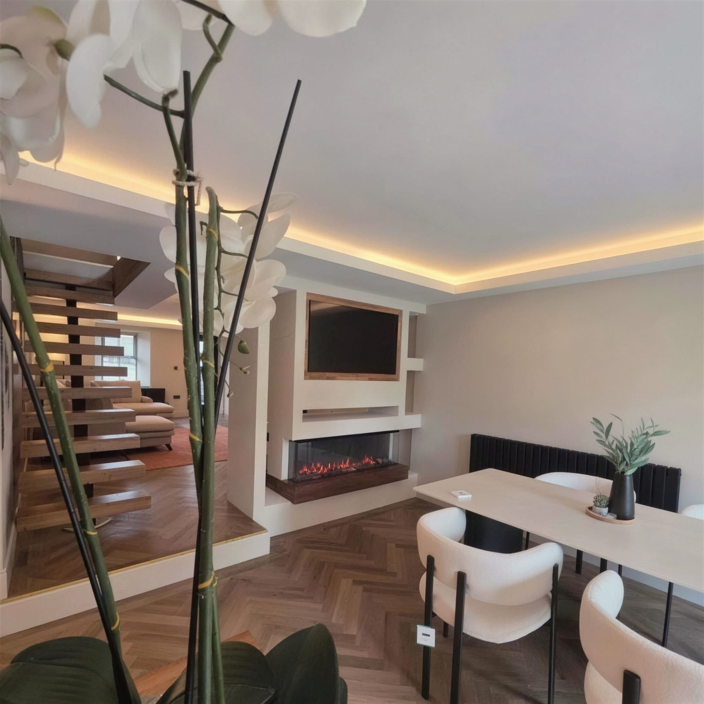 High-end open-plan interior with warm LED cove ceiling, plaster media wall with inset fire, herringbone floor and floating stairs, framed through a white orchid.