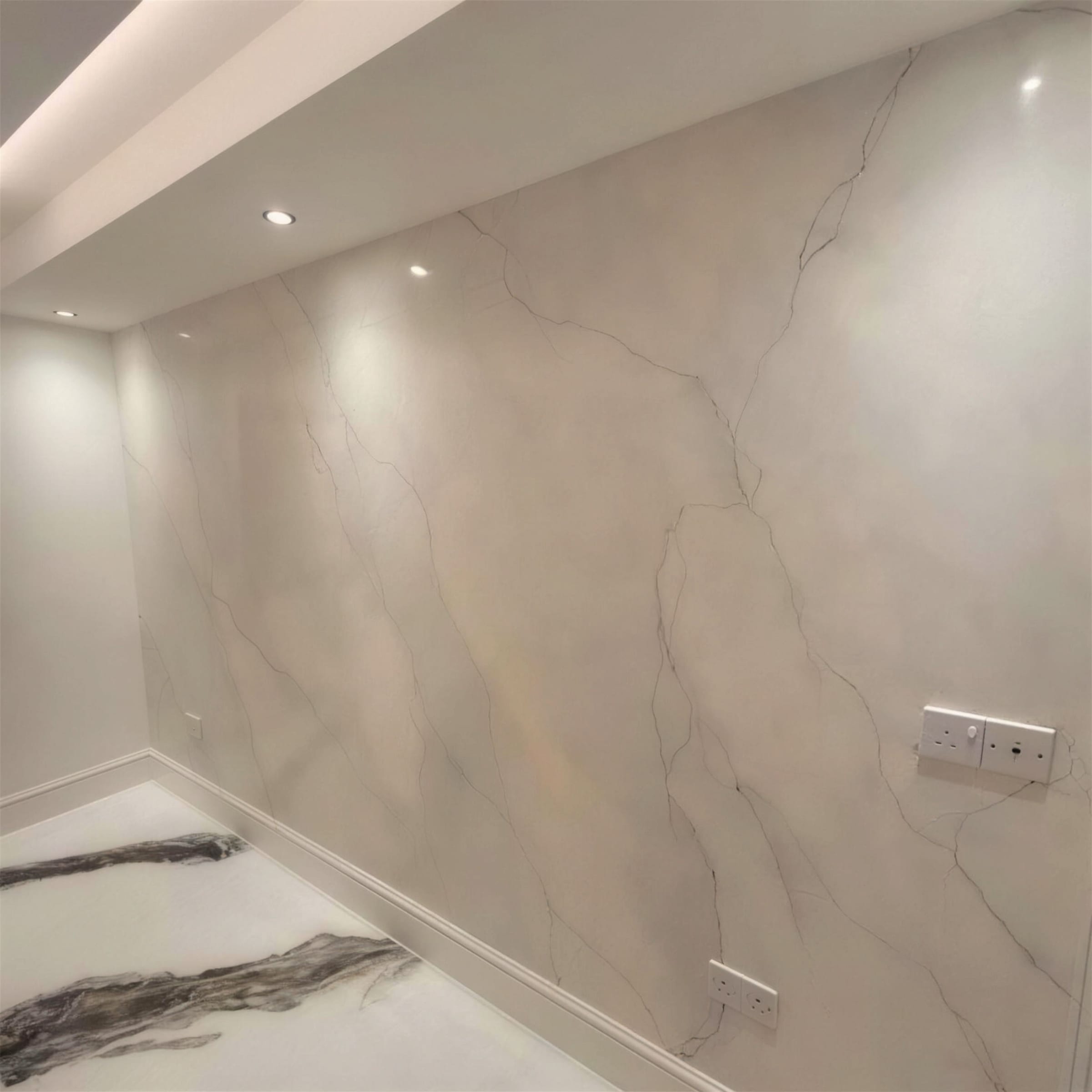 Full-height polished plaster wall with fine grey marble veining over warm ivory, lit by ceiling downlights above a book-matched marble floor.
