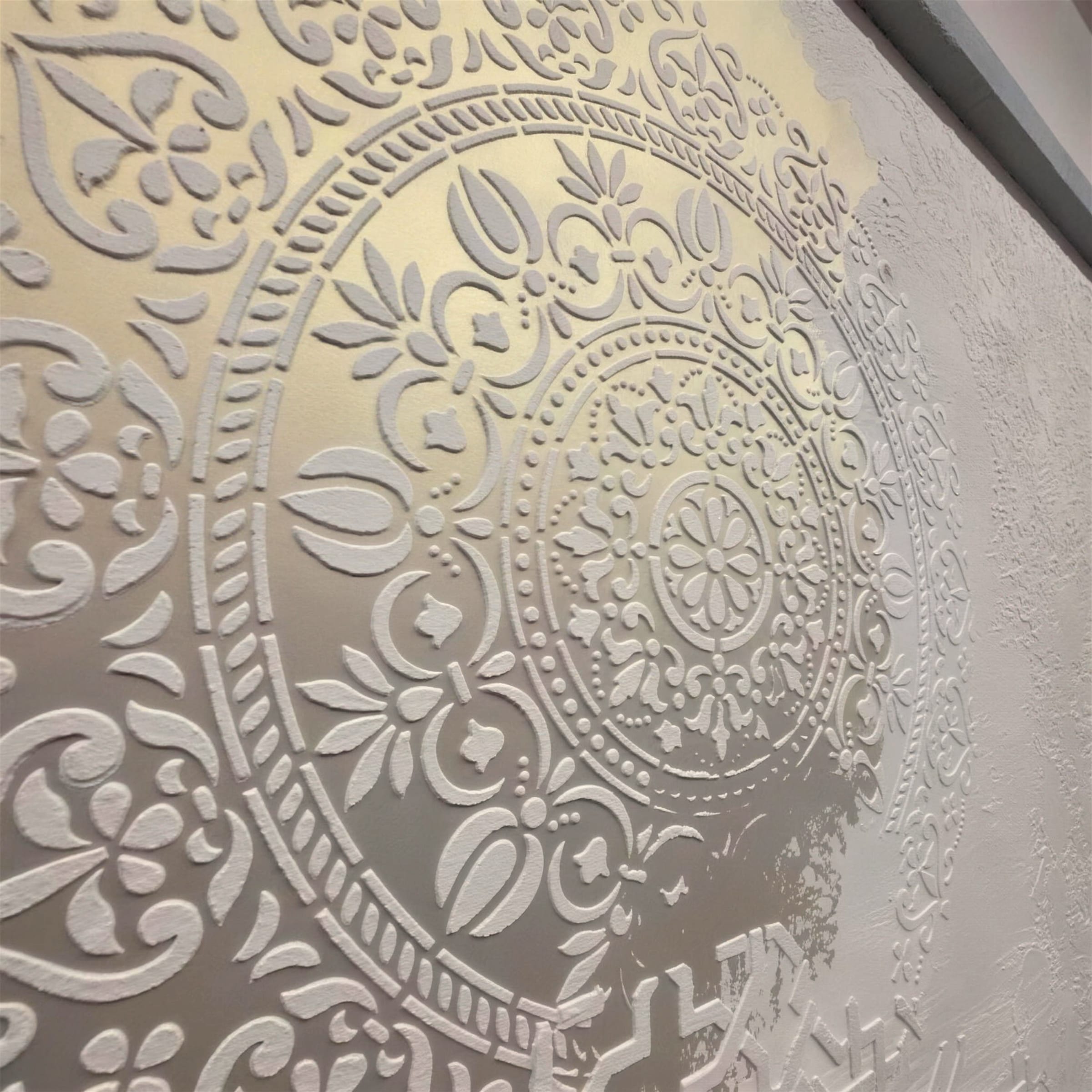 Close-up of an ornate raised mandala stencil in soft pearl and champagne tones, relief catching raking light across the plaster.