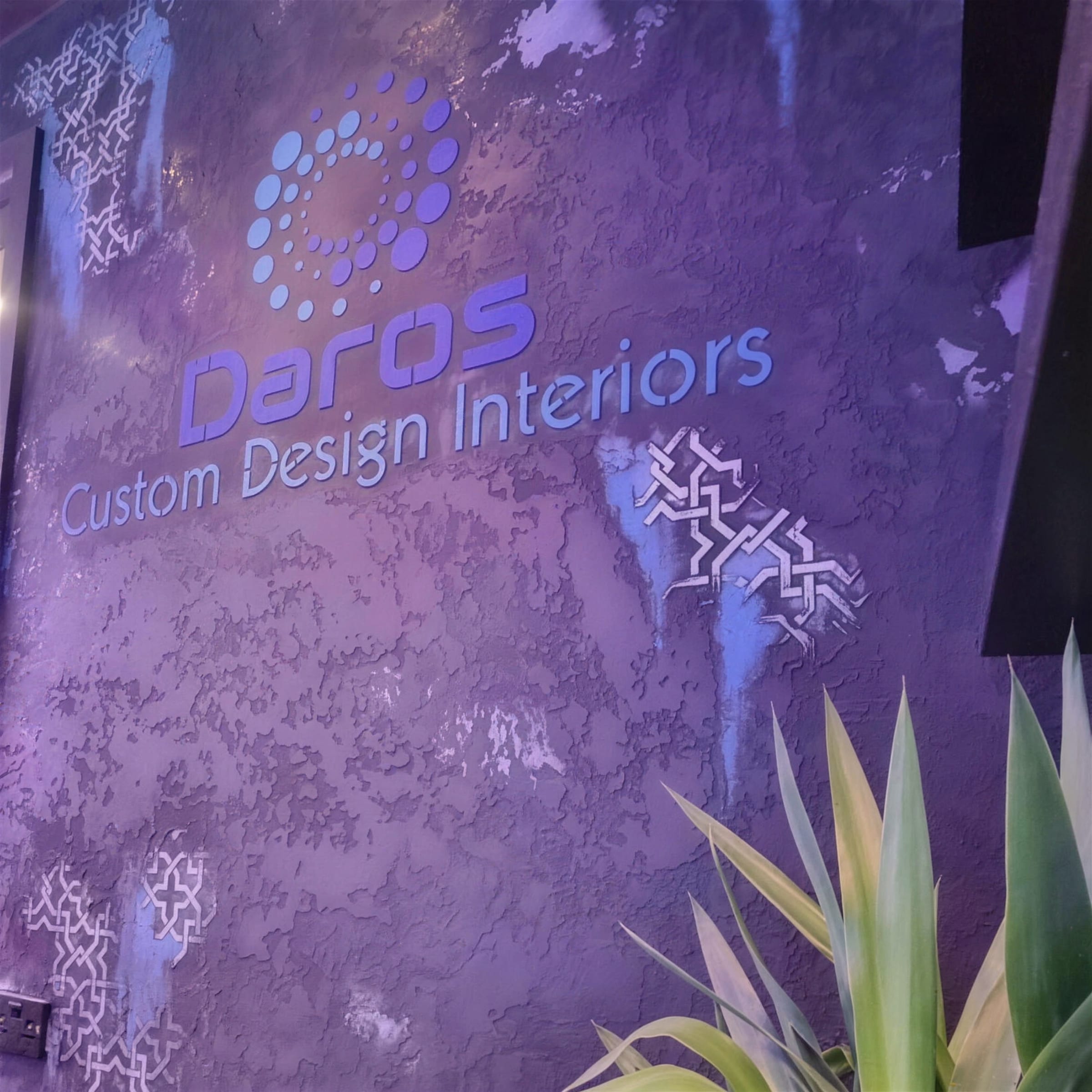 Distressed purple-bronze plaster wall with silver stencil accents and a backlit Daros Custom Design Interiors logo, plant in the foreground.