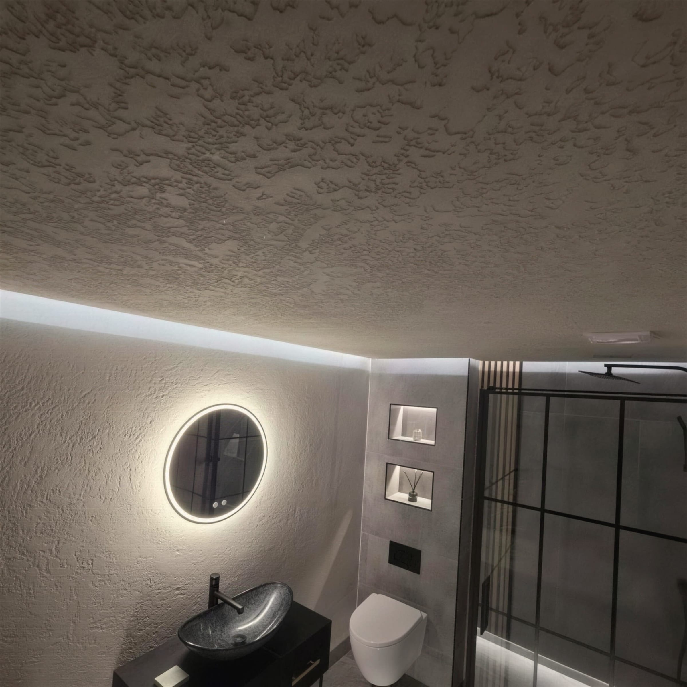 Moody bathroom with a stippled rustic plaster ceiling and walls, halo-lit round mirror, dark vessel basin and black-framed shower.