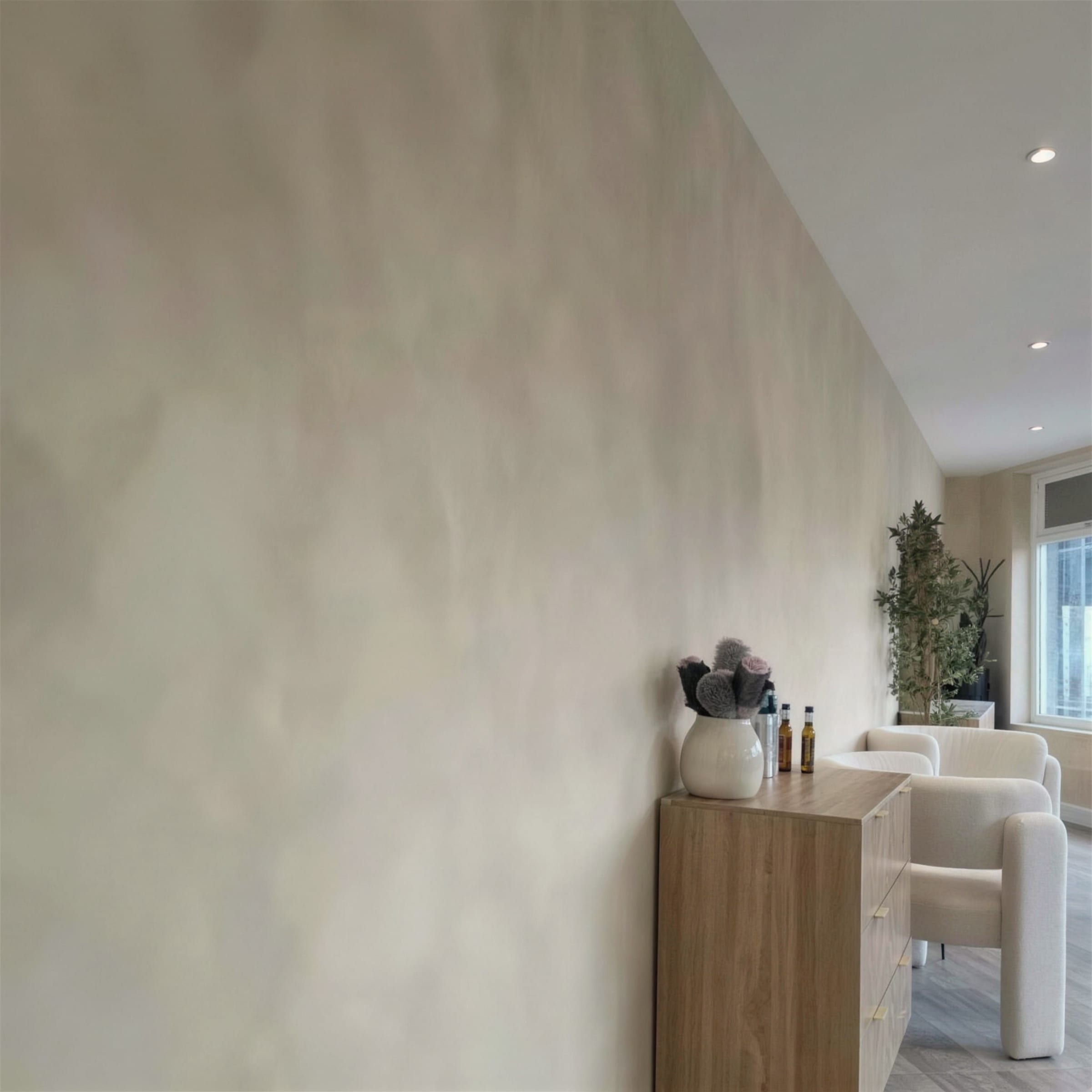 Raking perspective along a cloudy limewash plaster wall toward a styled seating corner with boucle chairs and greenery.